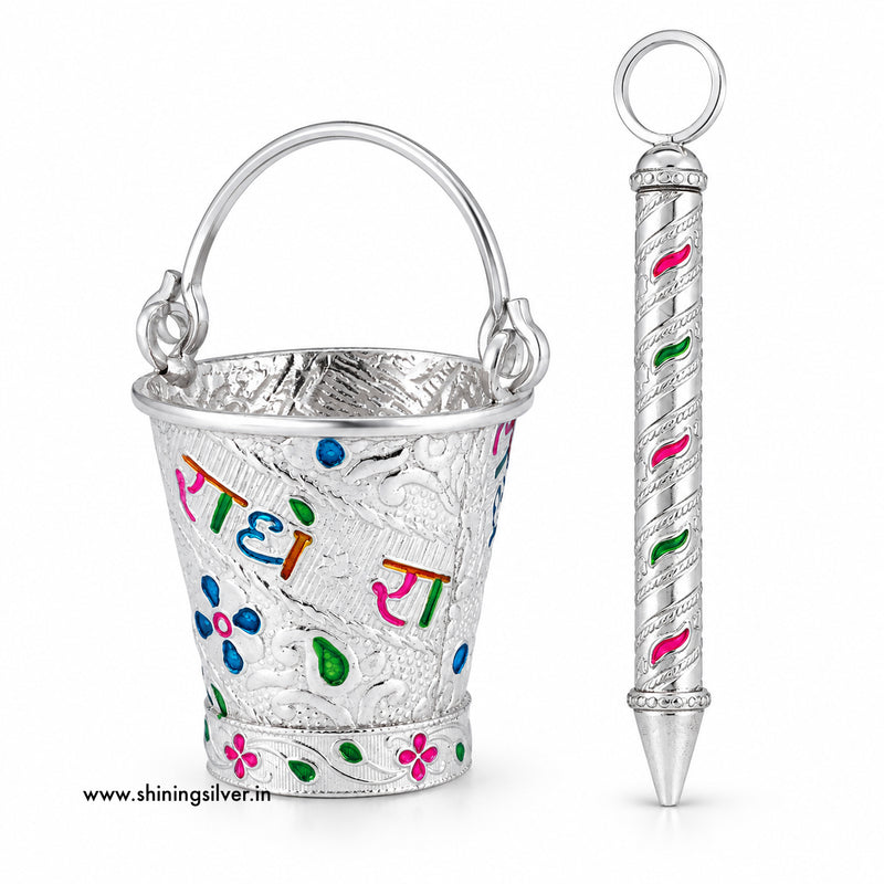 A silver-toned metal gift set featuring a small decorative bucket with a handle and a matching cylindrical pen or pointer, both decorated with colorful floral and script engravings. The bucket has the words 'Rati' in multicolored Devanagari script.