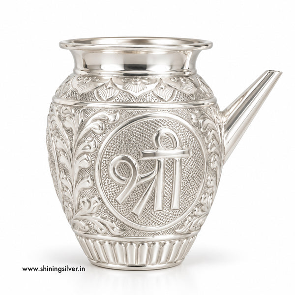 An ornate silver karwa with intricate floral engravings and a central circular emblem featuring the Shiva symbol, shown on a white background.