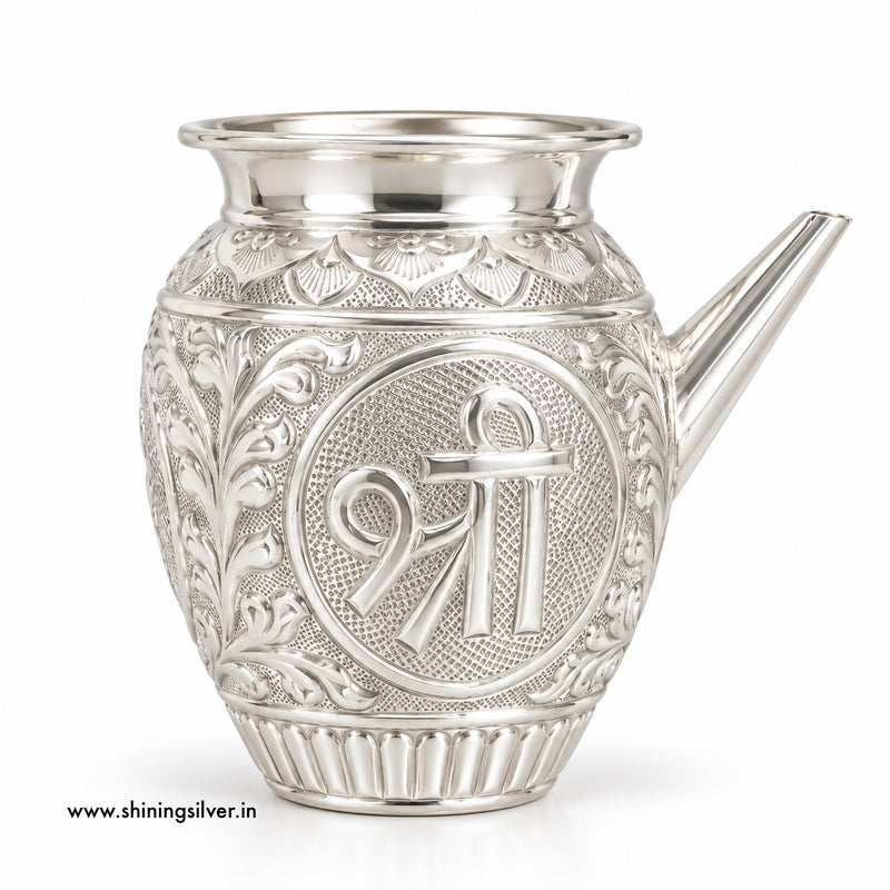 An ornate silver karwa with intricate floral engravings and a central circular emblem featuring the Shiva symbol, shown on a white background.