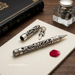 Decorative silver pen and clip on a piece of paper with an inkwell and book in the background. Shiningsilver.in