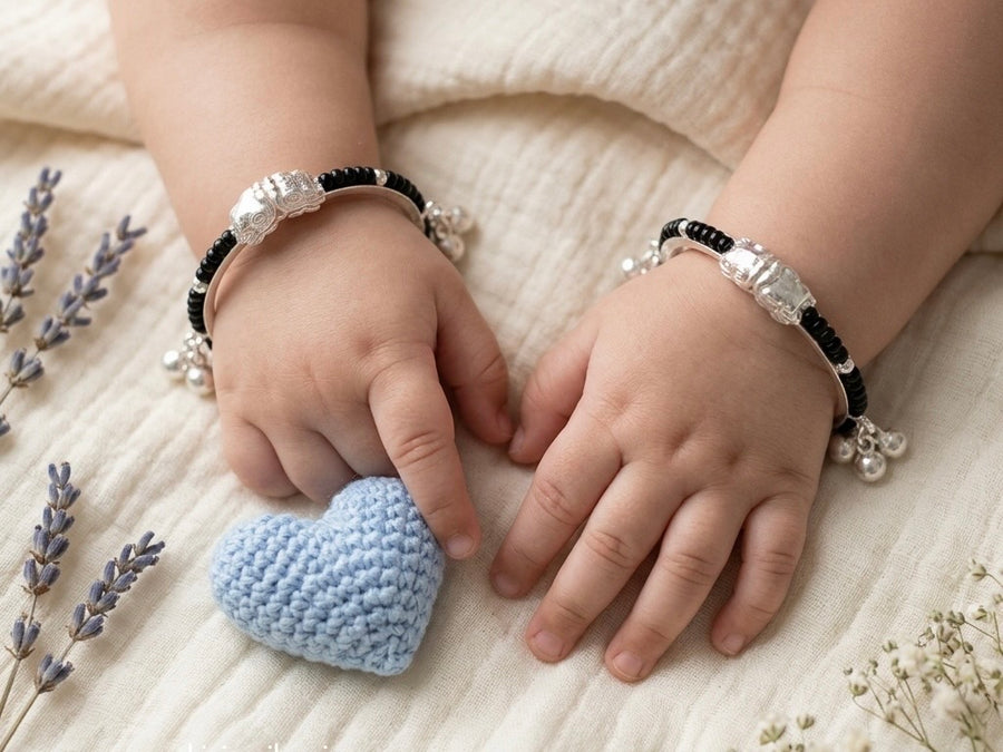 A pair of traditional Indian silver baby kadas featuring protective black beads, an intricate silver centerpiece, and hanging ghungroo bells.