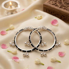 A pair of traditional Indian silver baby kadas featuring protective black beads, an intricate silver centerpiece, and hanging ghungroo bells.