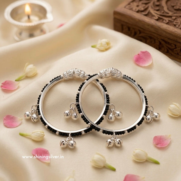 A pair of traditional Indian silver baby kadas featuring protective black beads, an intricate silver centerpiece, and hanging ghungroo bells.